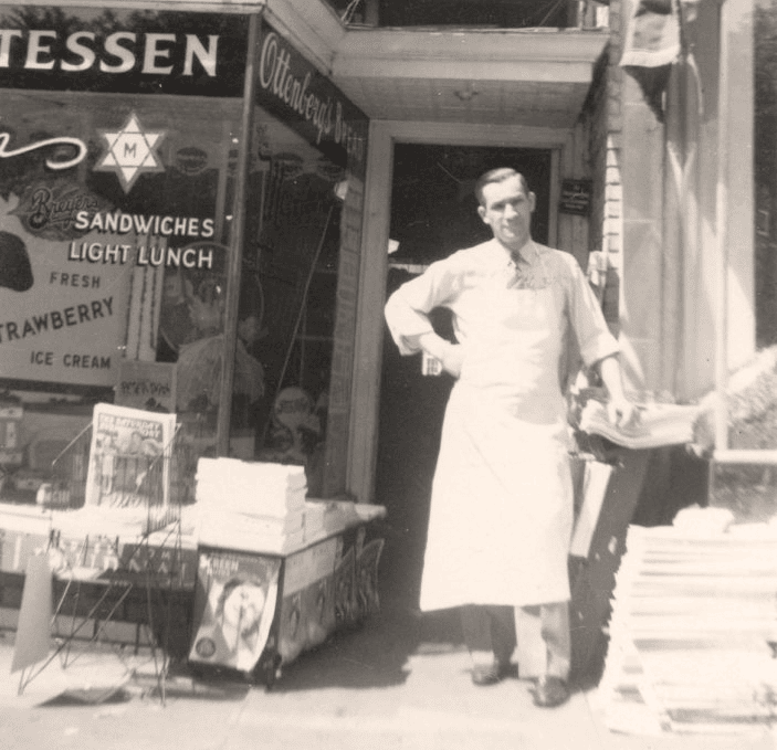 Vintage photograph of a deli man in a white apron standing in a shop doorway